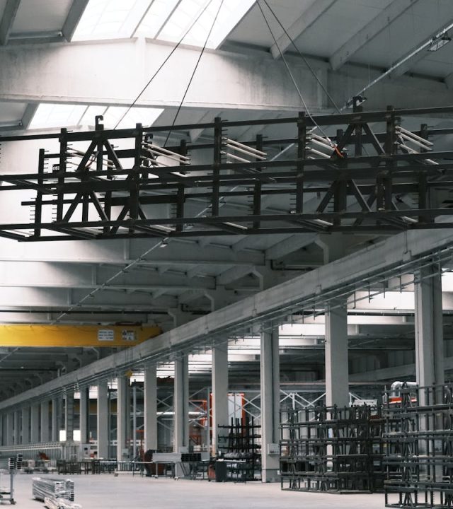 Spacious industrial warehouse interior with a yellow crane lifting metal framework.